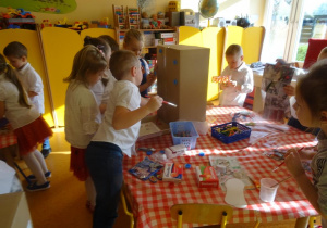 Grupa dzieci dekoruje karton papierowy kolorowymi ścinkami papieru.
